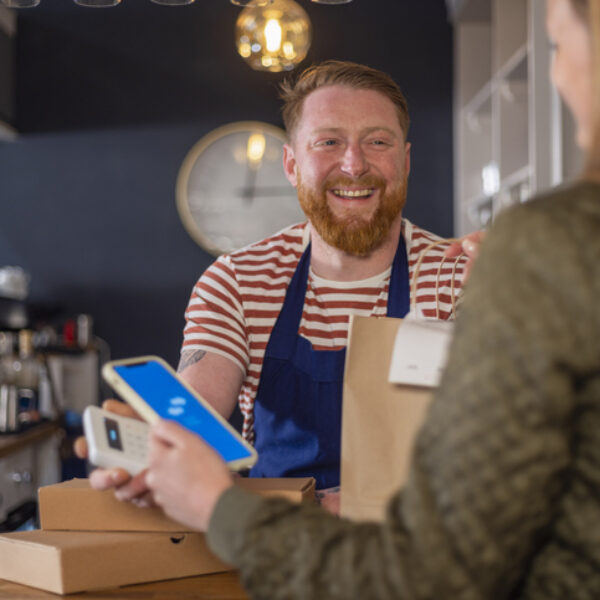 A shot of a mid-adult man wearing an apron, smiling and serving a customer at a restaurant in Northumberland, England. She has ordered food to take-out and she is collecting brown paper bags of food while she makes a contactless payment with her smart phone.